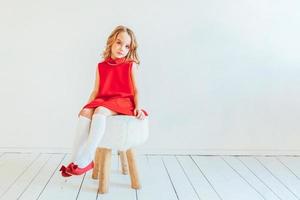 sucré peu fille dans rouge robe séance sur chaise contre blanc mur à maison, relaxant dans blanc brillant vivant pièce à l'intérieur. enfance écoliers jeunesse se détendre concept. photo