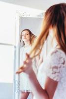 beauté maquillage Matin rutine l'amour toi même concept. Jeune adolescent fille à la recherche à réflexion dans miroir. Jeune positif femme portant blanc robe posant dans brillant lumière pièce contre blanc mur. photo