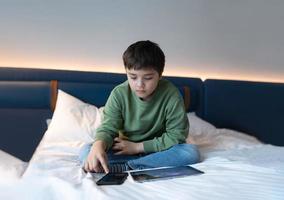 éducation concept, enfant en utilisant mobile téléphone en jouant Jeu sur l'Internet, homeschooling enfant Faire devoirs en ligne par tablette tampon à maison, garçon séance sur lit relaxant, en train de regarder dessin animé ou parlant avec ami photo