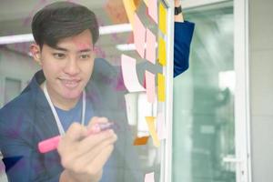 jeune homme d'affaires créatif tenant un marqueur et un plan d'écriture et partager une idée sur un mur de verre avec une note collante, un remue-méninges et discuter et formuler, des stratégies commerciales dans le bureau de démarrage technologique. photo