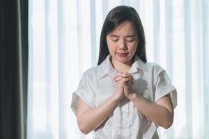 optimiste faire souhait, calme asiatique Jeune femme croyance, main dans prière, séance méditer sur canapé à maison, Christian fille prier à Dieu pour demander, demande avec fermé yeux rêver pour aider, Merci vous Dieu. photo