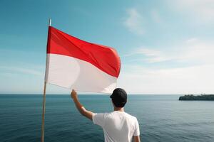 une homme en portant une rouge et blanc Indonésie drapeau à la recherche à le océan. ai généré photo