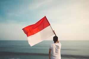 une homme en portant une rouge et blanc Indonésie drapeau à la recherche à le océan. ai généré photo