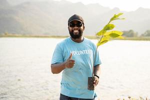 portrait de bénévole homme en portant pot avec vert plante souriant à caméra permanent sur rivière. protection de environnement et nature, écologie concept. photo