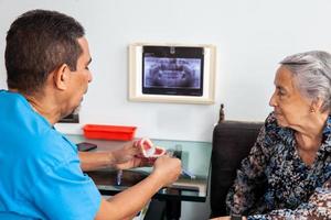 Masculin milieu -vieilli dentiste à le sien Bureau en utilisant une dentaire modèle à Explique traitement à le sien Sénior femme patient photo