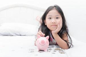 mignonne asiatique fille en mettant pièce de monnaie à porcin banque sur lit. photo