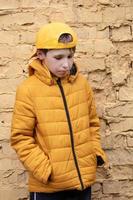 une enfant sur le rue. une en surpoids garçon est permanent contre une brique mur dans Jaune vêtements et une base-ball casquette. photo