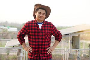 Beau asiatique homme porte chapeau, rouge plaid chemise, supporter à côté de solaire panneau à agriculture atterrir, mettre mains sur tailles. se sent confiant. concept, agriculteur installée photovoltaïque pour utilisation dans agriculture. photo