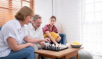 dans le vivant pièce de le loger, un personnes âgées couple est assis et se détend. à commencer en jouant échecs ensemble avec une échecs planche avec une fille applaudissement à côté de lui photo