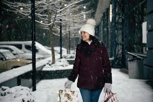 délicieux Multi-éthnique femme dans chaud vêtements, avec achats Sacs, en marchant vers le bas le ville rue, sur une neigeux hiver nuit photo