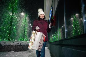 charmant femme avec achats Sacs et Noël cadeaux, en marchant vers le bas le rue, allégé par de fête éclairage photo