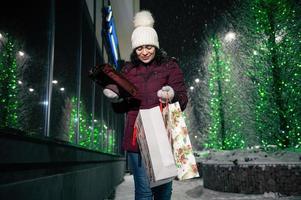 femme avec achats Sacs et Noël cadeaux, en marchant vers le bas le rue, allégé par éclairage à hiver neigeux nuit. photo