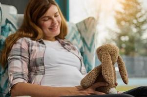 délicieux Enceinte femme est assis avec une peluche lapin, rêves de futur enfant à Accueil près fenêtre surplombant jardin. mignonne femme dans anticipation de bébé jouit sa insouciant grossesse et maternité laisser photo