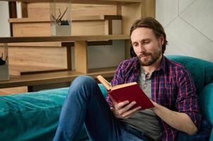 portrait de une Beau charmant barbu milieu vieilli caucasien homme en train de lire livre, séance sur canapé à Accueil intérieur, profiter une fin de semaine dans calme environnement photo