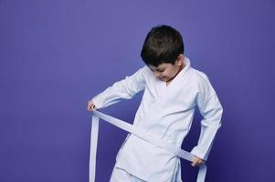 sur de soi content enfant garçon cravates blanc ceinture de aikido kimono autour taille. violet arrière-plan, copie espace. Oriental combat sport, martial les arts concept photo