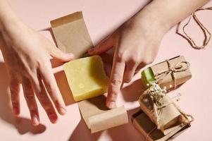 Haut vue de femme mains emballage Fait main biologique éco savon bar dans artisanat papier, sur isolé rose Contexte. photo