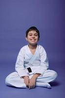 européen adolescent - aikido combattant - dans blanc kimono séance dans lotus pose tandis que pratiquant Oriental martial les arts sur violet Contexte avec copie espace photo