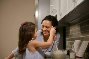 adorable peu fille ayant amusant, mélange Ingrédients et pétrissage pâte dans une verre bol tandis que sa aimant content mère enseignement sa à cuisinier crêpe pâte. rétrécir Mardi concept photo