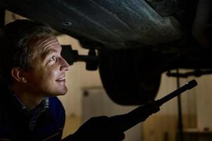 technicien vérification moderne voiture à voiture réparation magasin. auto mécanicien en utilisant une lampe de poche lampe pour inspecter le soubassement de le levé voiture dans une voiture service. voiture entretien concept photo