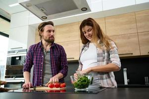 charmant Enceinte Jeune femme en train de préparer légume salade avec sa mari dans Accueil cuisine. caucasien hétérosexuel couple prendre plaisir unité tandis que cuisine en bonne santé aliments. national vie, maternité concept photo