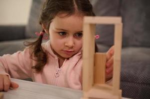 fermer portrait de adorable enfant, préscolaire fille dans rose sweat-shirt concentré sur bâtiment en bois structure avec en bois blocs. planche Jeux et bien moteur compétences développement concept photo