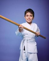 sur de soi fort concentré garçon - aikido lutteur dans blanc kimono des stands avec une en bois jo arme dans le sien mains. Oriental martial les arts entraine toi concept photo