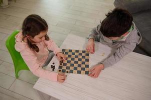 aérien vue de deux adorable caucasien élémentaire vieilli enfants, garçon et fille, frère et sœur ayant génial temps en jouant dames planche Jeu ensemble à Accueil intérieur. photo