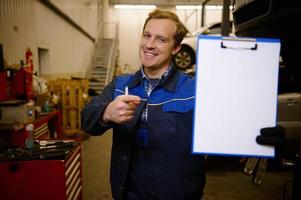 souriant caucasien auto mécanicien, voiture ingénieur, technicien montrer du doigt Vide presse-papiers feuille dans main avec copie espace pour voiture réparation un d liste de contrôle tandis que permanent contre voiture ascenseurs dans le réparation magasin garage photo