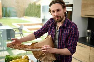 attrayant Beau Jeune caucasien homme déballage le éco papier sac avec en bonne santé nourriture et montrant le entier grain pain ou fraîchement cuit baguette, à la recherche à caméra permanent par cuisine table à Accueil photo