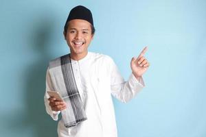 portrait de Jeune asiatique musulman homme en portant mobile téléphone avec souriant expression sur visage tandis que montrer du doigt doigt à le côté. isolé image sur bleu Contexte photo