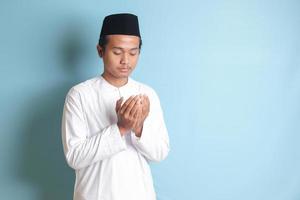 portrait de asiatique musulman homme dans blanc koko chemise avec calotte prier sérieusement avec le sien mains soulevé. isolé image sur bleu Contexte photo
