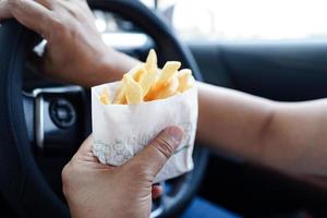 asiatique femme chauffeur tenir et manger français frites dans voiture, dangereux et risque un accident. photo