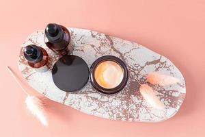 Haut vue de un ouvert pot de crème et deux cosmétique bouteilles de soins auto-administrés sérum sur une décoratif ovale plateau. beige Contexte. photo