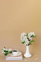 romantique printemps encore la vie sur une beige Contexte. une vase avec blanc Pomme fleurs, une tasse de thé dans une blanc porcelaine tasse et livres photo