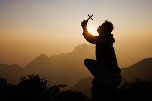 silhouette de une homme avec une crucifix dans le sien mains prier à Dieu. tandis que le Soleil est en hausse louange le religion. suprême Foi dans le Dieu de succès. aventure, voyage, religieux foi. photo