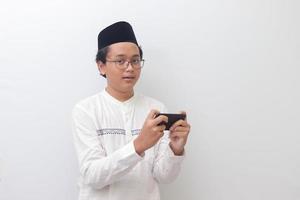 portrait de Jeune asiatique musulman homme élevage le sien poing, célébrer gagnant Jeu ou avoir bien nouvelles sur le sien mobile téléphone. isolé image sur blanc Contexte photo