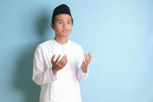 portrait de asiatique musulman homme dans blanc koko chemise avec calotte prier sérieusement avec le sien mains soulevé. isolé image sur bleu Contexte photo