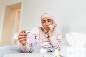 maladie, saisonnier virus problème concept. arabe femme étant malade ayant grippe mensonge sur lit à la recherche à Température sur thermomètre. malade femme avec hijab mensonge dans lit avec haute fièvre. photo