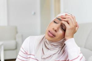 portrait de un arabe musulman femme séance sur une canapé à Accueil avec une mal de tête, sentiment douleur et avec un expression de étant indisposé. dérangé femme mensonge sur canapé sentiment fort mal de crâne migraine. photo