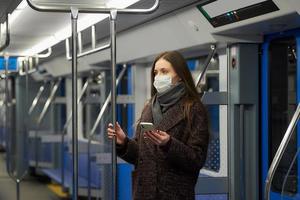 Une femme dans un masque facial est debout et à l'aide d'un smartphone dans une voiture de métro moderne