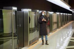 une femme dans un masque médical attend un train et tenant un smartphone