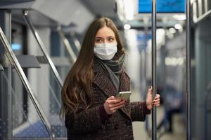 Une femme dans un masque facial est debout et à l'aide d'un smartphone dans une voiture de métro moderne
