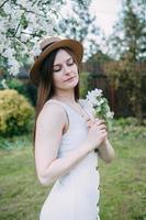 magnifique Jeune fille dans blanc robe et chapeau dans épanouissement Pomme verger. épanouissement Pomme des arbres avec blanc fleurs. photo