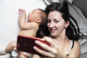 fermer de une mère et sa nouveau née bébé prise une selfie ou vidéo appel à leur père ou les proches dans lit. photo