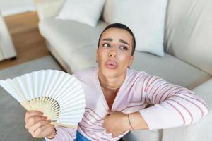 surchauffé femme asseoir sur canapé à Accueil ressentir chaud agitant avec main ventilateur refroidissement bas, transpiration fille se détendre sur canapé dans vivant pièce tenir vaciller souffrir de chaleur, non air Conditionneur système photo