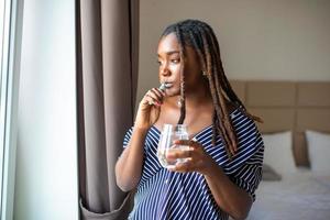 Jeune africain femme brossage les dents avec brosse à dents, en portant verre de l'eau et à la recherche dans fenêtre photo