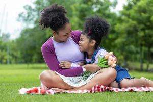 africain mère en portant bouquet de tulipe fleurs et le fille étreindre chaque autre tandis que Heureusement profiter pique-nique dans le Publique parc pendant été pour famille l'amour et se soucier dans de la mère journée fête photo