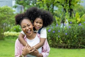 une mère afro-américaine joue sur le dos et s'embrasse avec sa jeune fille tout en faisant un pique-nique d'été dans le parc public pour le concept de bien-être et de bonheur photo