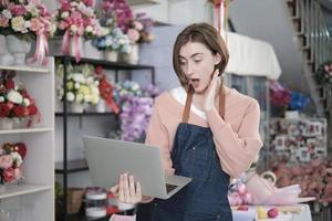 un Jeune magnifique blanc femelle fleuriste propriétaire surpris et sous le choc achat commande profit, en ligne le revenu travail dans portable à brillant fleur magasin magasin, petit commerce électronique entreprise, content pme entrepreneur. photo