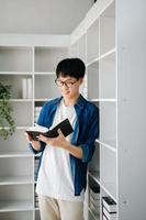 Masculin étudiant prise Remarques de une livre à bibliothèque, Jeune asiatique séance à bureau Faire affectations dans Université bibliothèque photo